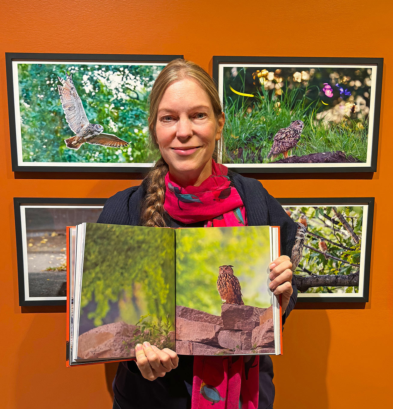 Photographer Anke Frohlich is holding The Book of Flaco, opened to a page with a double spread of one of her photographs. She is standing in front of three of her images on display at the exhibit. 